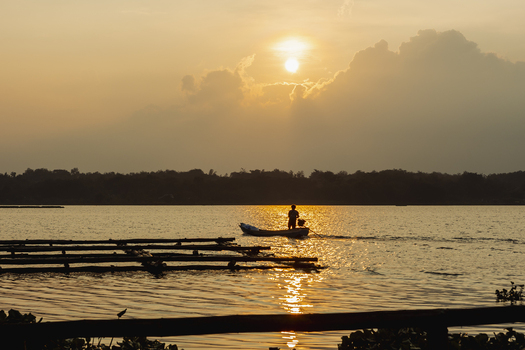 View of a fisherman in a small boat on a calm lake during a golden sunset with wooden structures in the foreground Kecamatan Tawangmangu Jawa Tengah Indonesia.