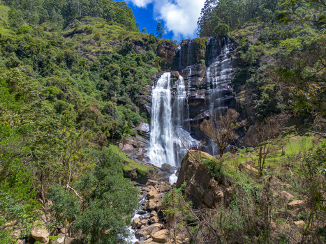 Aerial view of Bomburu Ella Waterfall cascading down rocky cliffs surrounded by lush green forest with people standing on the rock ledge in Nuwara Eliya Central Province Sri Lanka. Aerial view of Bomburu Ella Waterfall cascading down rocky cliffs surrounded by lush green forest with people standing on the rock ledge in Nuwara Eliya Central Province Sri Lanka.