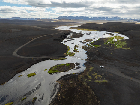 Aerial view of a winding river flowing through a vast dark volcanic landscape with patches of green moss and a small vehicle on a dirt road in Iceland. Aerial view of a winding river flowing through a vast dark volcanic landscape with patches of green moss and a small vehicle on a dirt road in Iceland.