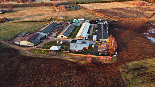 Aerial view of a rural farm complex with multiple long agricultural buildings surrounding plowed fields and dirt paths under the warm light of the golden hour in Latvia. Aerial view of a rural farm complex with multiple long agricultural buildings surrounding plowed fields and dirt paths under the warm light of the golden hour in Latvia.