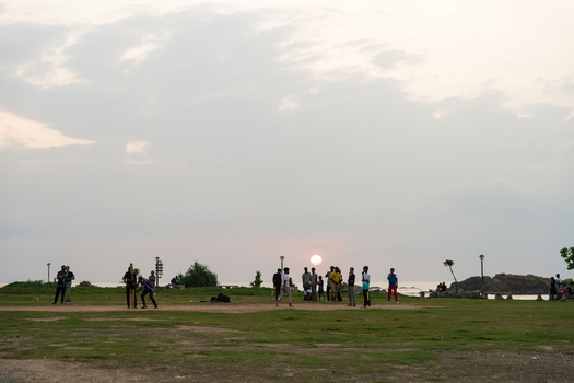 Sri Lanka 15 January 2026 View of people playing cricket on a grassy field at sunset with the ocean and rocky coastline in the background.
