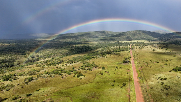 Aerial view of a vibrant double rainbow arching over rolling green hills and a red dirt road under a stormy sky in Waterberg District Municipality Limpopo South Africa. Aerial view of a vibrant double rainbow arching over rolling green hills and a red dirt road under a stormy sky in Waterberg District Municipality Limpopo South Africa.