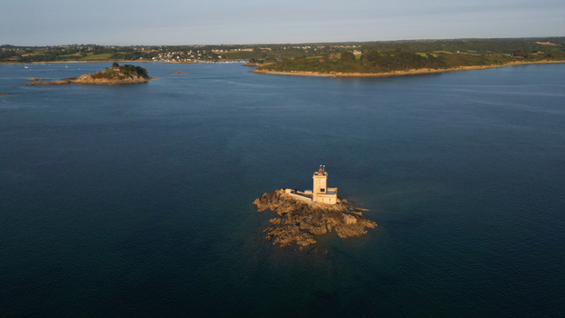 Aerial view of a lighthouse rising from ile Noire islet surrounded by deep blue waters with the distant coastline in the background Plouezoc h Finistere France.