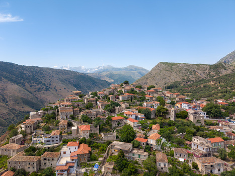 Aerial view of the historic hillside village with traditional stone houses and red tiled roofs surrounded by rugged mountains in Qeparo Fshat Vlore County Albania. Aerial view of the historic hillside village with traditional stone houses and red tiled roofs surrounded by rugged mountains in Qeparo Fshat Vlore County Albania.