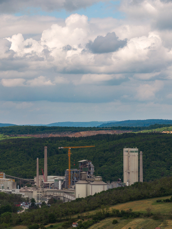 Aerial view of an industrial plant with tall chimneys silos and a yellow crane nestled among lush green forests and rolling hills in Sivice South Moravian Region Czechia.