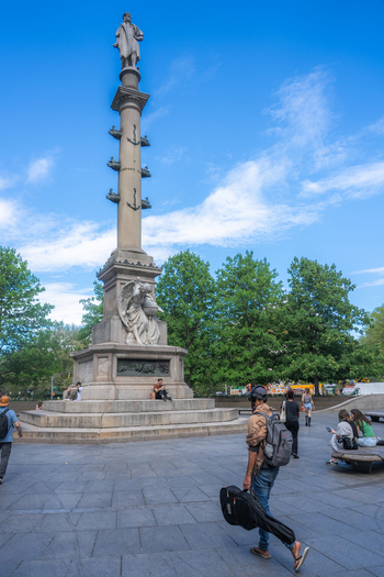 View of the Columbus Monument at Columbus Circle with people walking and lush green trees under a blue sky in New York New York United States.