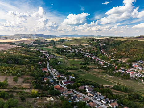 Aerial view of Csabdi village Hungary with homes and winding roads among wooded hills patchwork farmland and a broad rural valley under cumulus clouds.