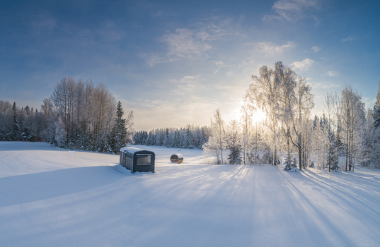 Aerial view of a small cabin and a barrel sauna set in a vast snow covered field surrounded by frost covered trees under a bright winter sun in Latvia. Aerial view of a small cabin and a barrel sauna set in a vast snow covered field surrounded by frost covered trees under a bright winter sun in Latvia.
