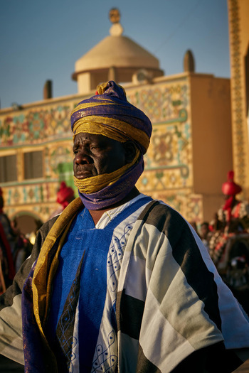 Zaria city Nigeria 22 March 2026 View of a man in vibrant blue and yellow traditional attire with a solemn gaze standing before an intricately patterned building with a golden dome. Zaria city Nigeria 22 March 2026 View of a man in vibrant blue and yellow traditional attire with a solemn gaze standing before an intricately patterned building with a golden dome.