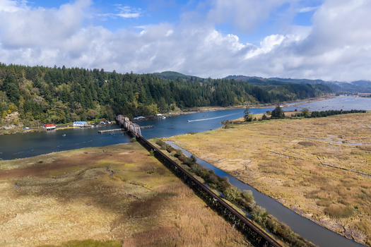 Aerial view of the Siuslaw River Train Bridge spanning the river surrounded by lush wetlands dense forests and a cloudy blue sky in Florence Oregon United States.