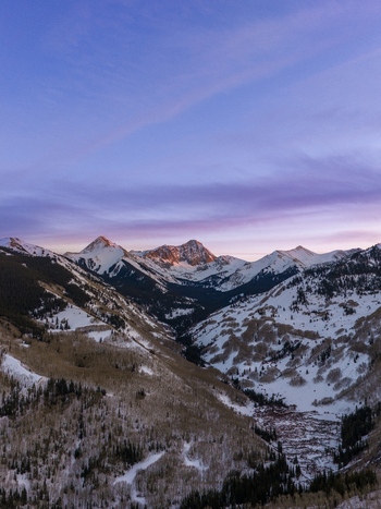 Aerial view of Capitol Peak in the Rocky Mountains at winter sunset featuring snow covered slopes evergreen forests and a vibrant purple and orange sky in Aspen Colorado United States.