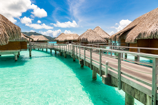 View of overwater bungalows with thatched roofs connected by wooden walkways over the clear turquoise lagoon in Bora Bora Leeward Islands French Polynesia.