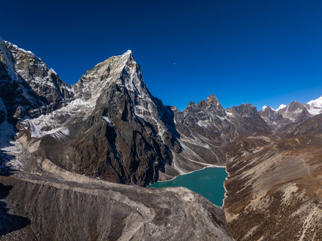 Aerial view of the dramatic Himalayan mountain peaks rising above a turquoise glacial lake with rugged rocky slopes and snow capped summits under a clear blue sky in Nepal.