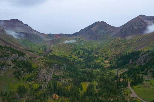 Aerial view of the lush Yankee Boy Basin with Mt. Sneffels in the background featuring dense coniferous forests rocky slopes and a winding dirt road in Ouray Colorado United States.