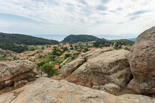 View of Adatepe village nestled among rocky hills and lush greenery with a distant sea view under a cloudy sky in Adatepe Canakkale Turkey. View of Adatepe village nestled among rocky hills and lush greenery with a distant sea view under a cloudy sky in Adatepe Canakkale Turkey.