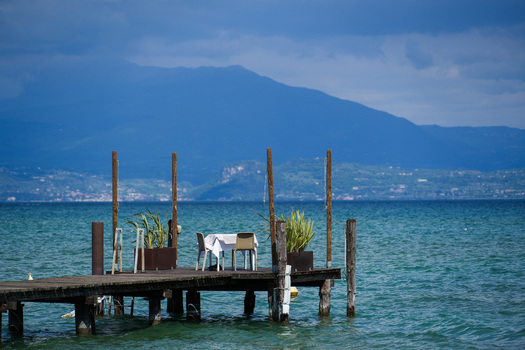 View of a table for two set on a wooden pier overlooking the blue waters of Lake Garda with mountains in the background Sirmione Lombardy Italy.