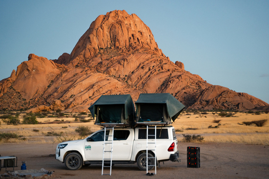 Spitzkoppe Namibia 12 March 2026 View of a white pickup truck fitted with rooftop tents stands silhouetted against the rugged ochre colored Spitzkoppe mountains.