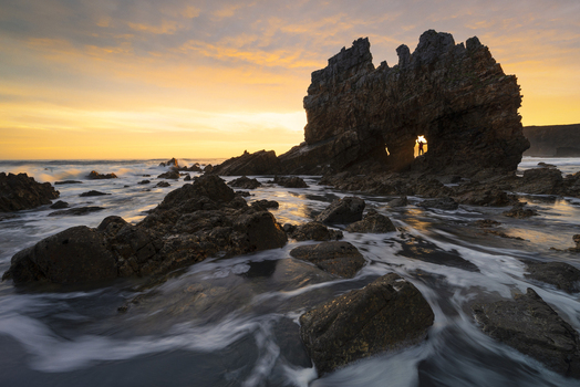 View of Playa de Portizuelo with a person standing in a rock arch during a golden sunset over the crashing waves of the Atlantic coast Spain. View of Playa de Portizuelo with a person standing in a rock arch during a golden sunset over the crashing waves of the Atlantic coast Spain.