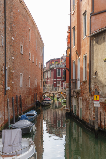 View of a narrow canal in Venice Italy lined with historic brick buildings and moored small boats with a small stone bridge in the distance Venice Veneto Italy. View of a narrow canal in Venice Italy lined with historic brick buildings and moored small boats with a small stone bridge in the distance Venice Veneto Italy.