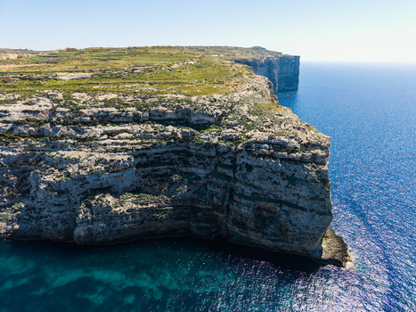 Aerial view of the rugged limestone cliffs of Dwejra Bay overlooking the turquoise Mediterranean Sea under a clear blue sky in Gozo Region Malta. Aerial view of the rugged limestone cliffs of Dwejra Bay overlooking the turquoise Mediterranean Sea under a clear blue sky in Gozo Region Malta.
