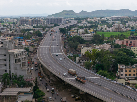 Aerial view of a highway flyover winding through the urban landscape with trucks and cars surrounded by buildings and distant hills in Karad Maharashtra India.