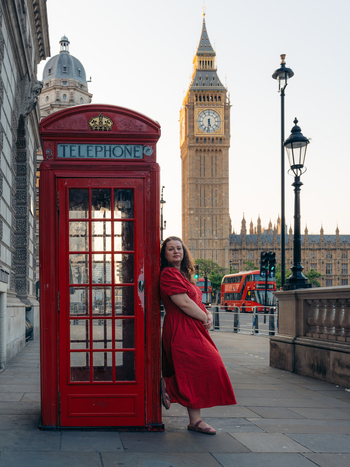 View of a woman in a red dress leans against a classic red telephone booth with Big Ben rising majestically in the background London England United Kingdom. View of a woman in a red dress leans against a classic red telephone booth with Big Ben rising majestically in the background London England United Kingdom.