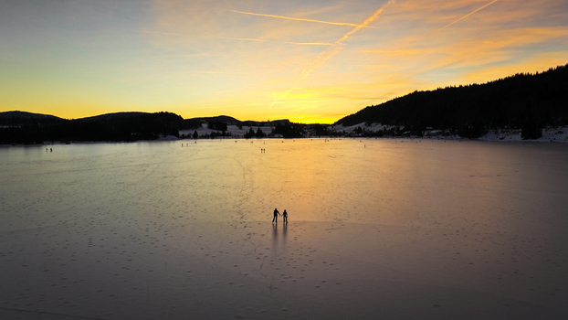 Aerial view of two figures skating across the vast frozen expanse of Lac des Rousses under the warm glow of the setting sun Les Rousses Bourgogne Franche Comté France. Aerial view of two figures skating across the vast frozen expanse of Lac des Rousses under the warm glow of the setting sun Les Rousses Bourgogne Franche Comté France.