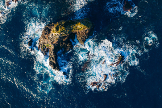 Aerial view of a rugged rocky islet surrounded by deep blue ocean water and white crashing waves in Ponta Delgada Acores Portugal. Aerial view of a rugged rocky islet surrounded by deep blue ocean water and white crashing waves in Ponta Delgada Acores Portugal.