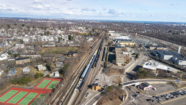 Aerial view of a train cutting through the urban sprawl alongside tennis courts and commercial buildings under a vast sky Braintree Massachusetts United States.