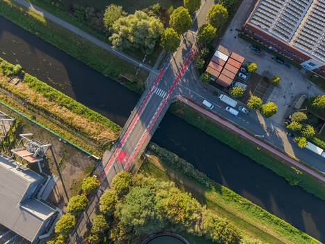 Aerial view of a bridge crossing a canal in an industrial area featuring red marked cycling lanes surrounding greenery and nearby warehouse buildings in Vilvoorde Flanders Belgium.