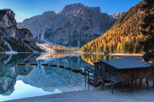 View of the wooden boathouse on the calm turquoise waters of Lago di Braies with the towering peaks of the Dolomites reflected in the lake and autumn trees along the shore in Italy.