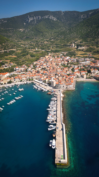 Aerial view of the coastal town of Komiza featuring its harbor long stone pier and red roofed houses nestled against green hills Komiza Split Dalmatia County Croatia.