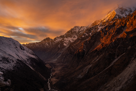 Aerial view of snow capped Himalayan peaks illuminated by the warm golden light of sunset with a deep valley and a winding river below in Nepal.