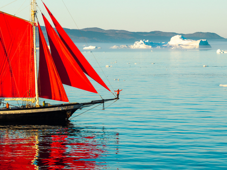 Greenland 06 July 2026 Aerial view of a sailboat with striking red sails navigating through icy waters with icebergs in the background and a person standing on the bowsprit. Greenland 06 July 2026 Aerial view of a sailboat with striking red sails navigating through icy waters with icebergs in the background and a person standing on the bowsprit.