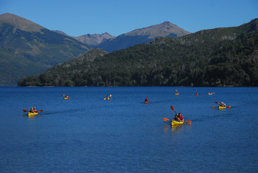 View of people paddling yellow and red kayaks on the blue waters of Nahuel Huapi Lake with forested hills and mountains in San Carlos de Bariloche Rio Negro Argentina. View of people paddling yellow and red kayaks on the blue waters of Nahuel Huapi Lake with forested hills and mountains in San Carlos de Bariloche Rio Negro Argentina.