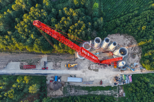 Aerial view of a large red crawler crane assembling wind turbine tower segments at a construction site surrounded by dense forest in Hofgeismar Hessen Germany. Aerial view of a large red crawler crane assembling wind turbine tower segments at a construction site surrounded by dense forest in Hofgeismar Hessen Germany.