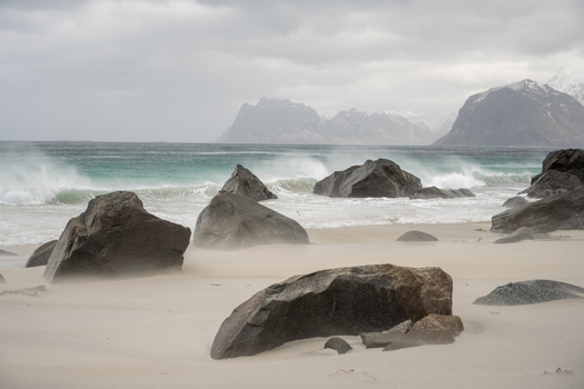 View of a sandy beach with large dark rocks crashing waves and distant mountains under an overcast sky in Leknes Lofoten Norway.