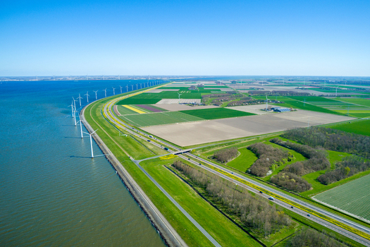 Aerial view of wind turbines lining the coast along a dike with agricultural fields and a road in the Netherlands.