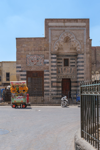 Cairo Egypt 20 April 2026 View of the Zawya and Sabil of Sultan Faraj Ibn Barquq featuring intricate Mamluk architecture striped stone walls and a traditional fruit cart on the street. Cairo Egypt 20 April 2026 View of the Zawya and Sabil of Sultan Faraj Ibn Barquq featuring intricate Mamluk architecture striped stone walls and a traditional fruit cart on the street.