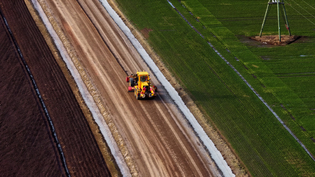 Aerial view of a yellow motor grader leveling a dirt road between a dark plowed field and a vibrant green field with a power line pylon in Latvia. Aerial view of a yellow motor grader leveling a dirt road between a dark plowed field and a vibrant green field with a power line pylon in Latvia.