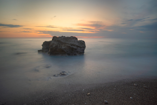 View of a solitary rock formation rises from the silky ethereal sea under a pastel sky painted with fading light and soft hues Kathisma Greece. View of a solitary rock formation rises from the silky ethereal sea under a pastel sky painted with fading light and soft hues Kathisma Greece.