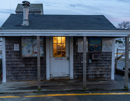 Edgartown United States 14 May 2026 View of a small wooden ferry ticket booth with shingle siding a lit window and ferry schedules posted on the exterior at dusk.