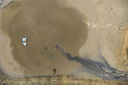 Aerial view of a small white boat floating in a shallow muddy pool of water with sediment patterns and a drainage pipe in Latvia.