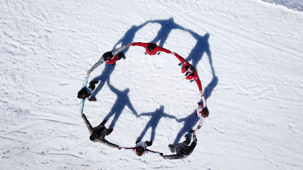 Aerial view of a group of people in winter gear holding hands in a circle on snow with long shadows cast on the white ground in Verbier Val de Bagnes Valais Switzerland.
