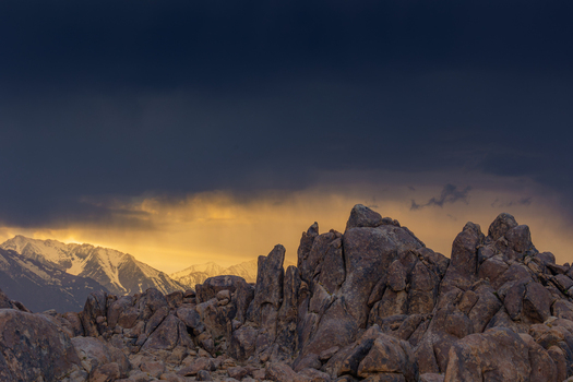 View of Death Valley National Park featuring rugged rock formations in the foreground and snow capped mountains under a dramatic stormy sky with golden sunset light in the United States.