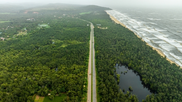Aerial view of a straight highway running through dense forest along the coastline with ocean waves hitting the beach in Honnavar Karnataka India.