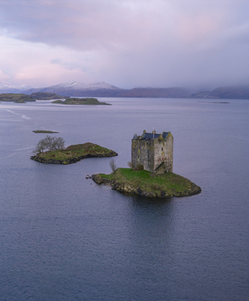 Aerial view of Castle Stalker on a small green island in the middle of a calm loch with snow capped mountains under a twilight sky in Appin Scotland United Kingdom. Aerial view of Castle Stalker on a small green island in the middle of a calm loch with snow capped mountains under a twilight sky in Appin Scotland United Kingdom.