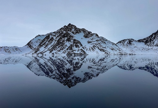 Aerial view of a snow covered mountain peak perfectly reflected in the calm waters of a fjord under an overcast sky in Tasiilaq Sermersooq Greenland. Aerial view of a snow covered mountain peak perfectly reflected in the calm waters of a fjord under an overcast sky in Tasiilaq Sermersooq Greenland.