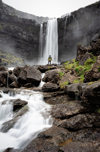 View of a person standing before a powerful waterfall cascading over dark wet rocks in a rugged mountainous landscape in the Faroe Islands.