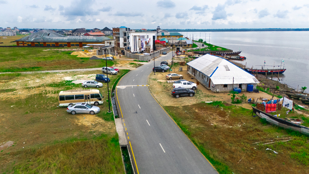 Aerial view of a coastal road winding through a small settlement with parked vehicles traditional wooden boats moored along the shoreline and modern buildings in Minima Rivers Nigeria.